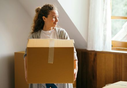 Young smiling lady in casual outfit with big cardboard box in hands standing and looking away near bed in modern bedroom of newly purchased house